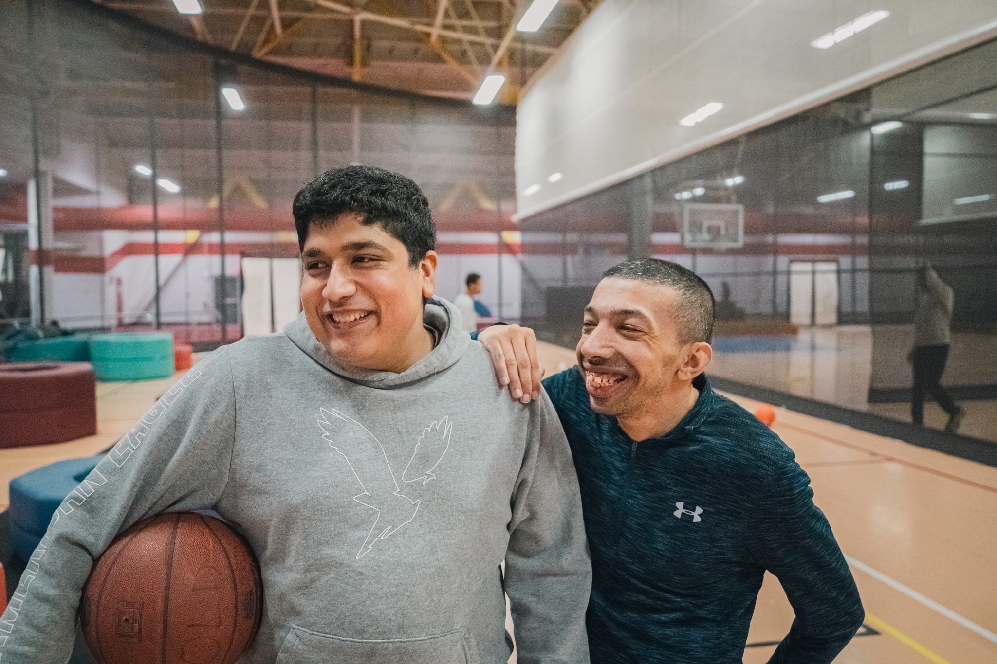 Two basketball players smile together on an indoor court, with one holding a basketball.