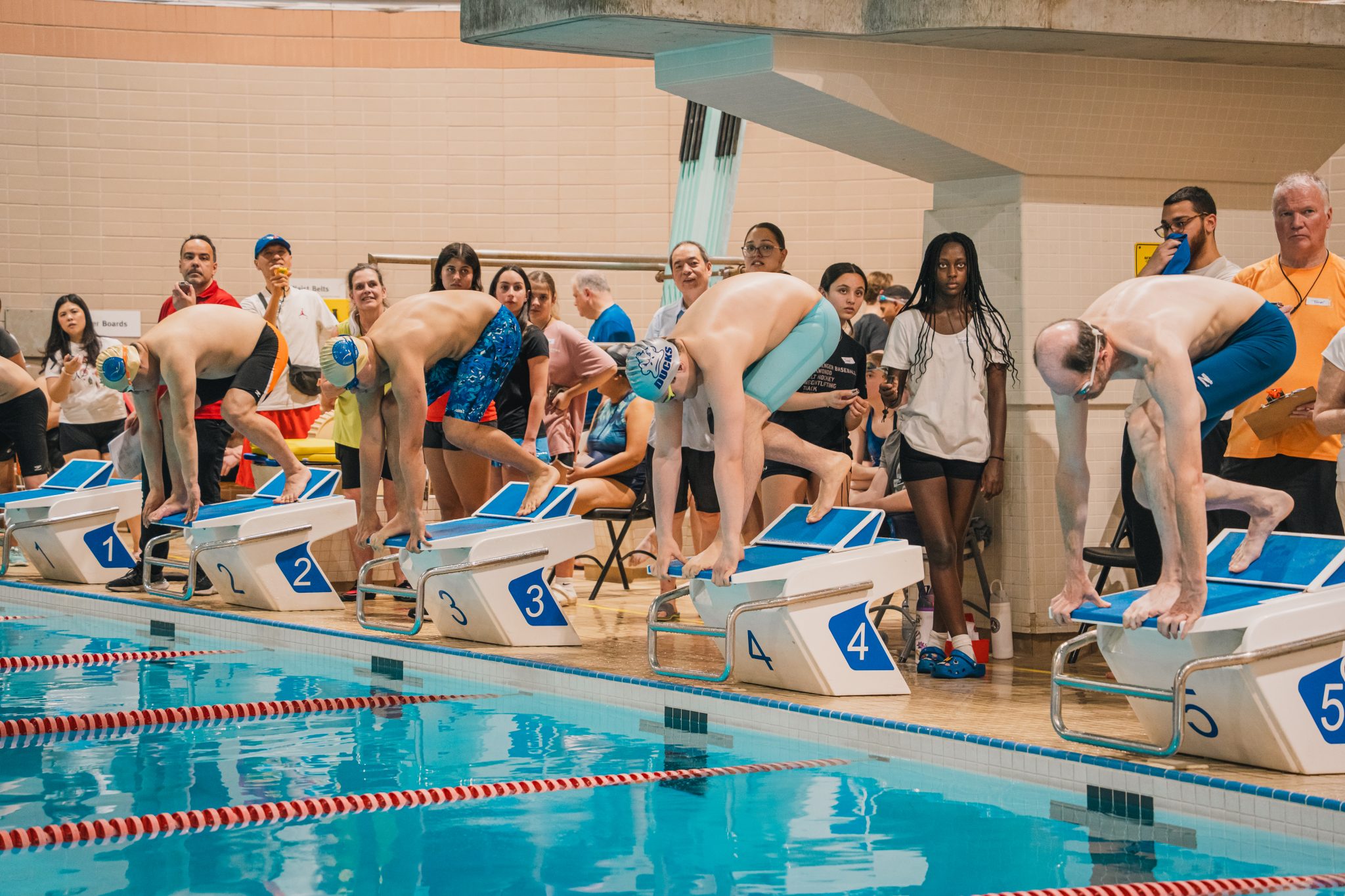 Swimmers prepare on starting blocks at the edge of an indoor pool before the start of a race as spectators watch from the deck.