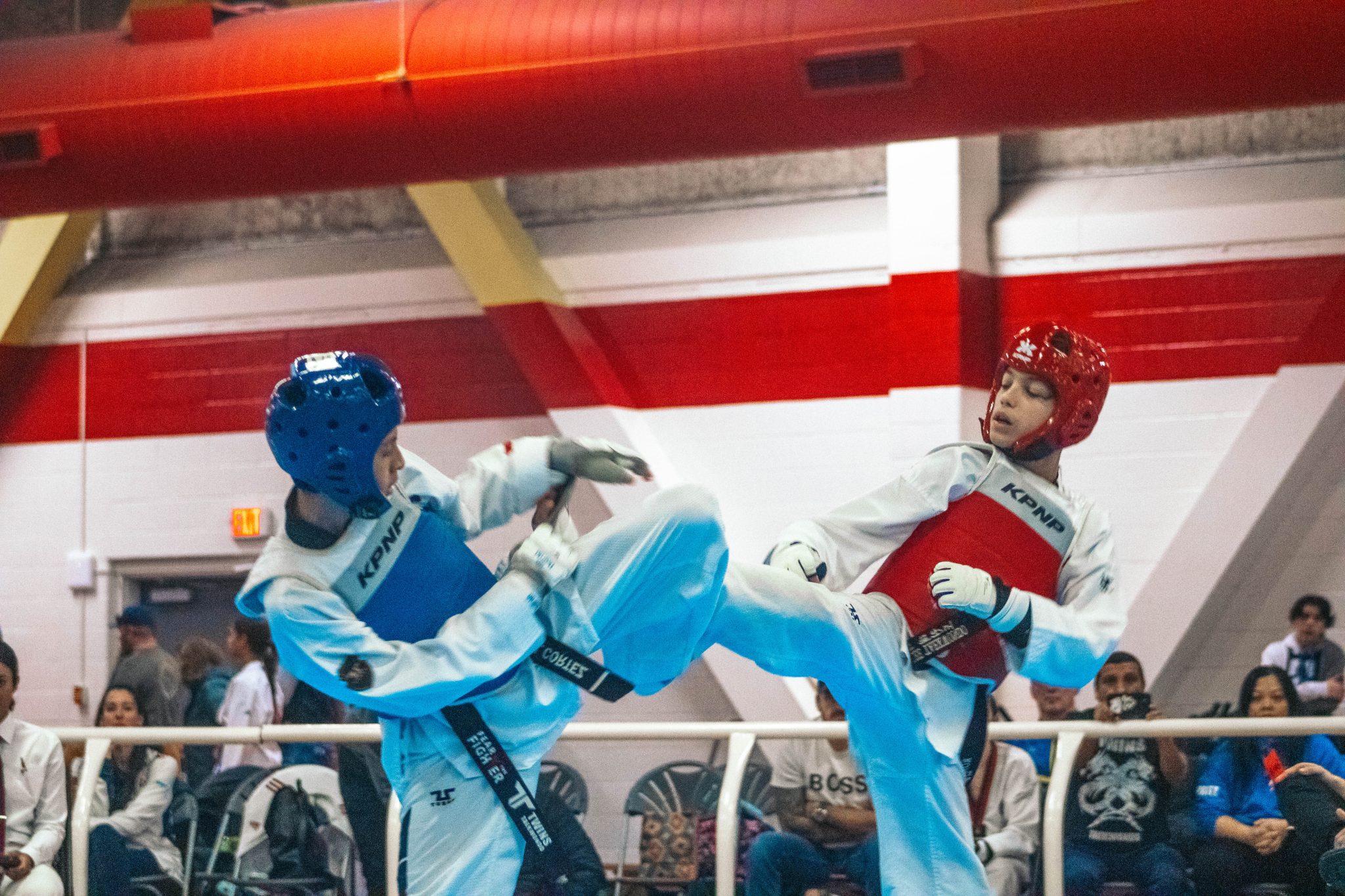 Two taekwondo athletes wearing protective headgear and chest protectors exchange simultaneous kicks during a sparring match inside a gymnasium. One athlete in blue gear and one in red gear are both airborne as spectators watch from behind a barrier, capturing the intensity and timing of the match.
