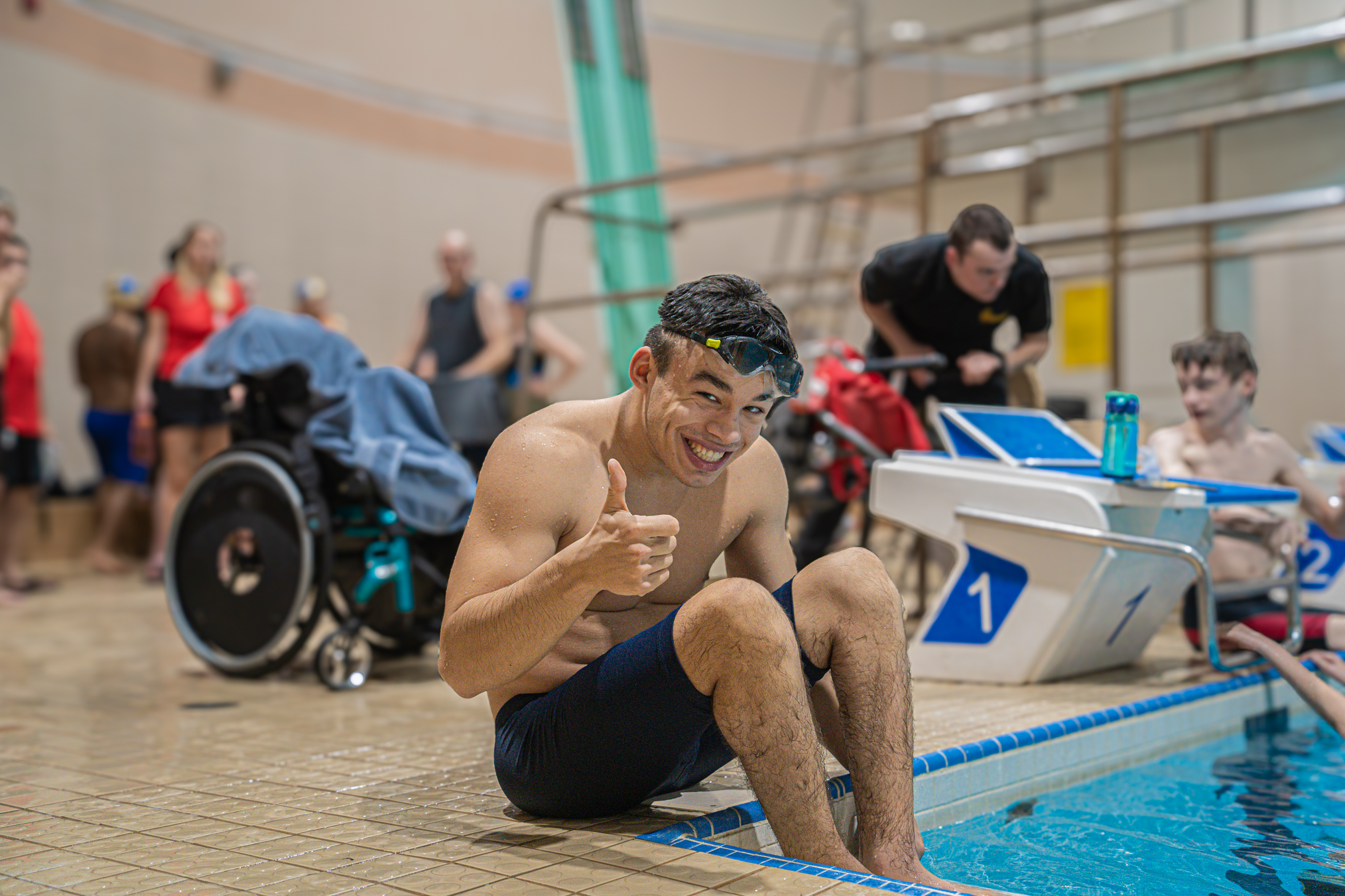 Smiling swimmer sits on the pool deck giving a thumbs up before or after a swim meet, with teammates and coaches in the background.