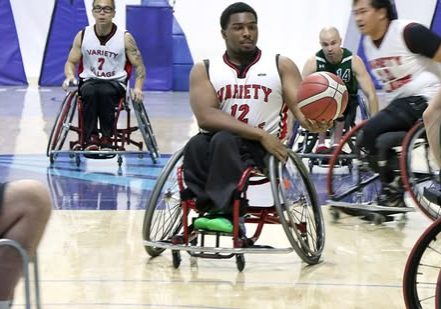 Wheelchair basketball player dribbles the ball up the court during a game while teammates and opponents race into position.