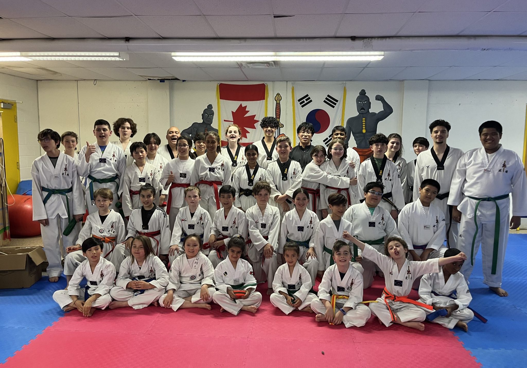 A group of karate athletes and coaches pose together in a dojo wearing white uniforms and coloured belts after a training session.