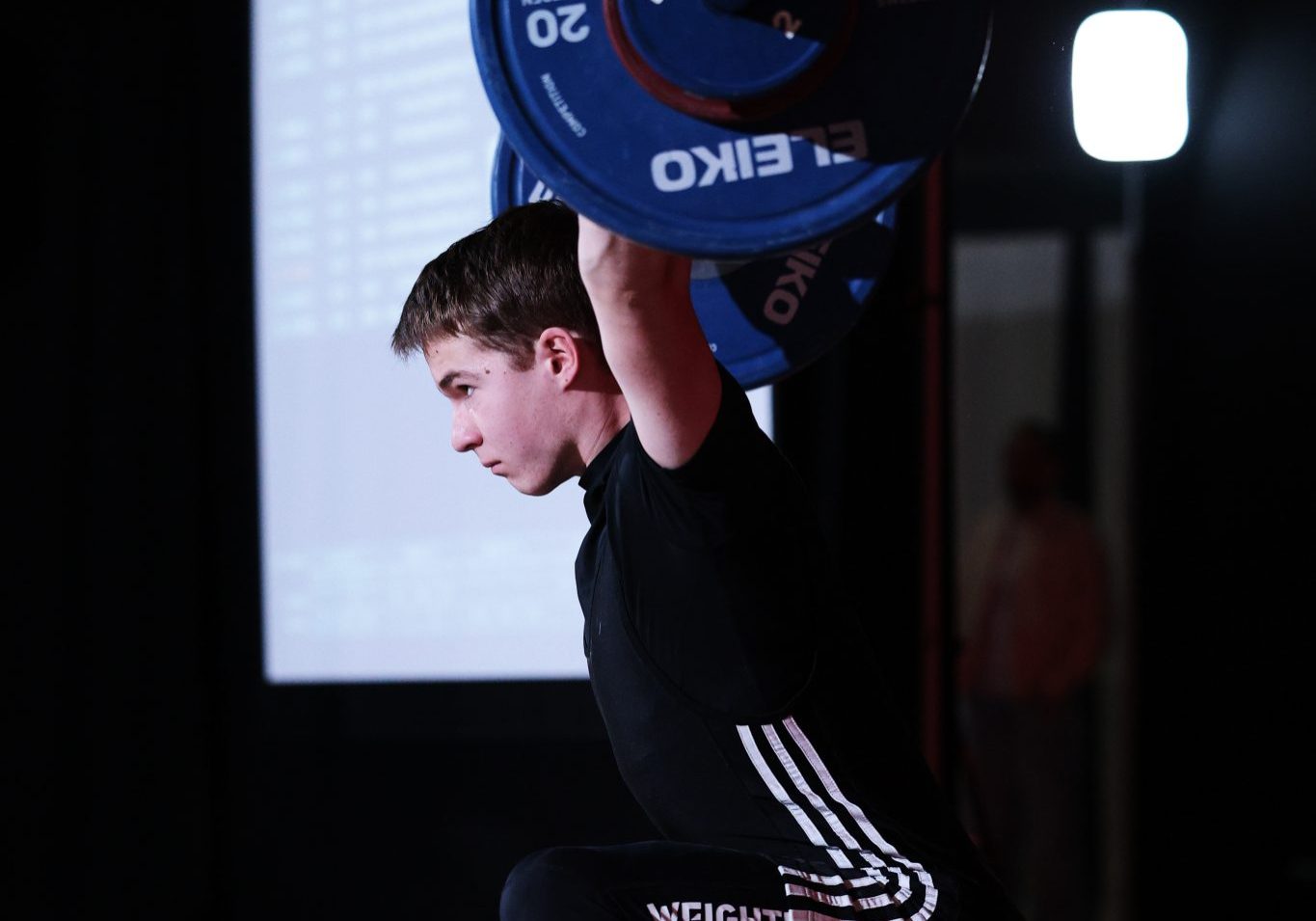 A young athlete performs an overhead squat with a loaded Olympic barbell during a weightlifting competition. Standing on a lifting platform under bright lights, the athlete is captured in a deep squat with the bar stabilized overhead, while a scoreboard screen is visible in the background.