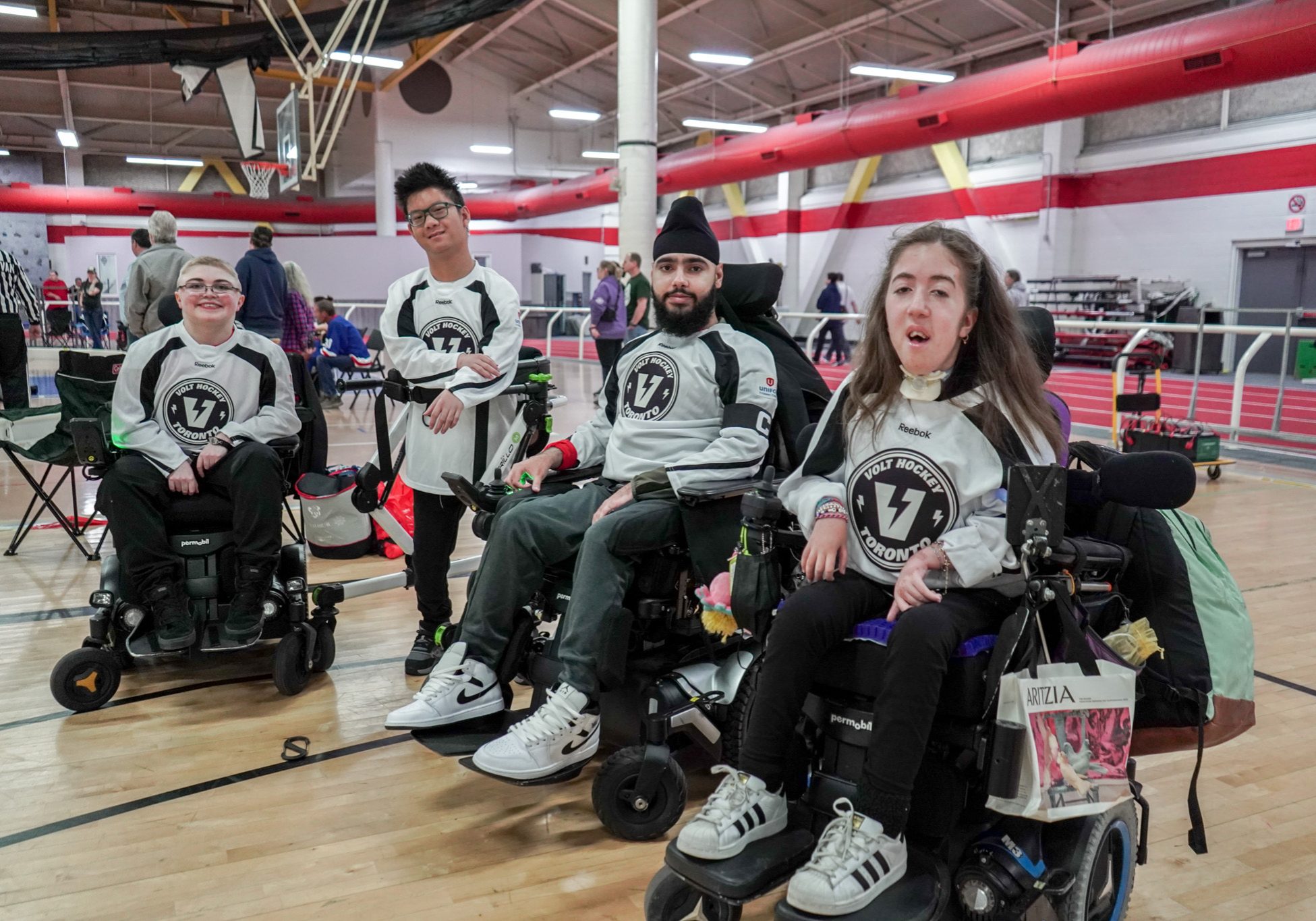 Four powerchair hockey athletes pose together on an indoor court wearing team jerseys.