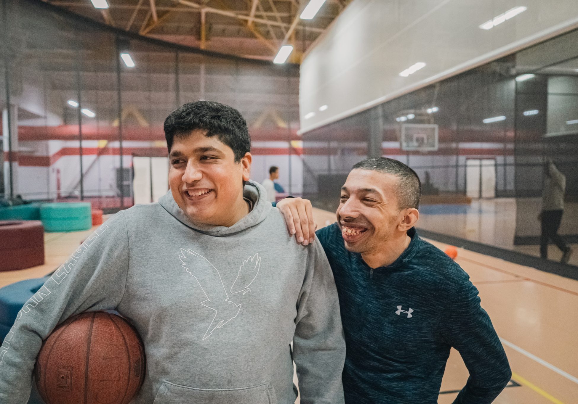 Two basketball players smile together on an indoor court, with one holding a basketball.