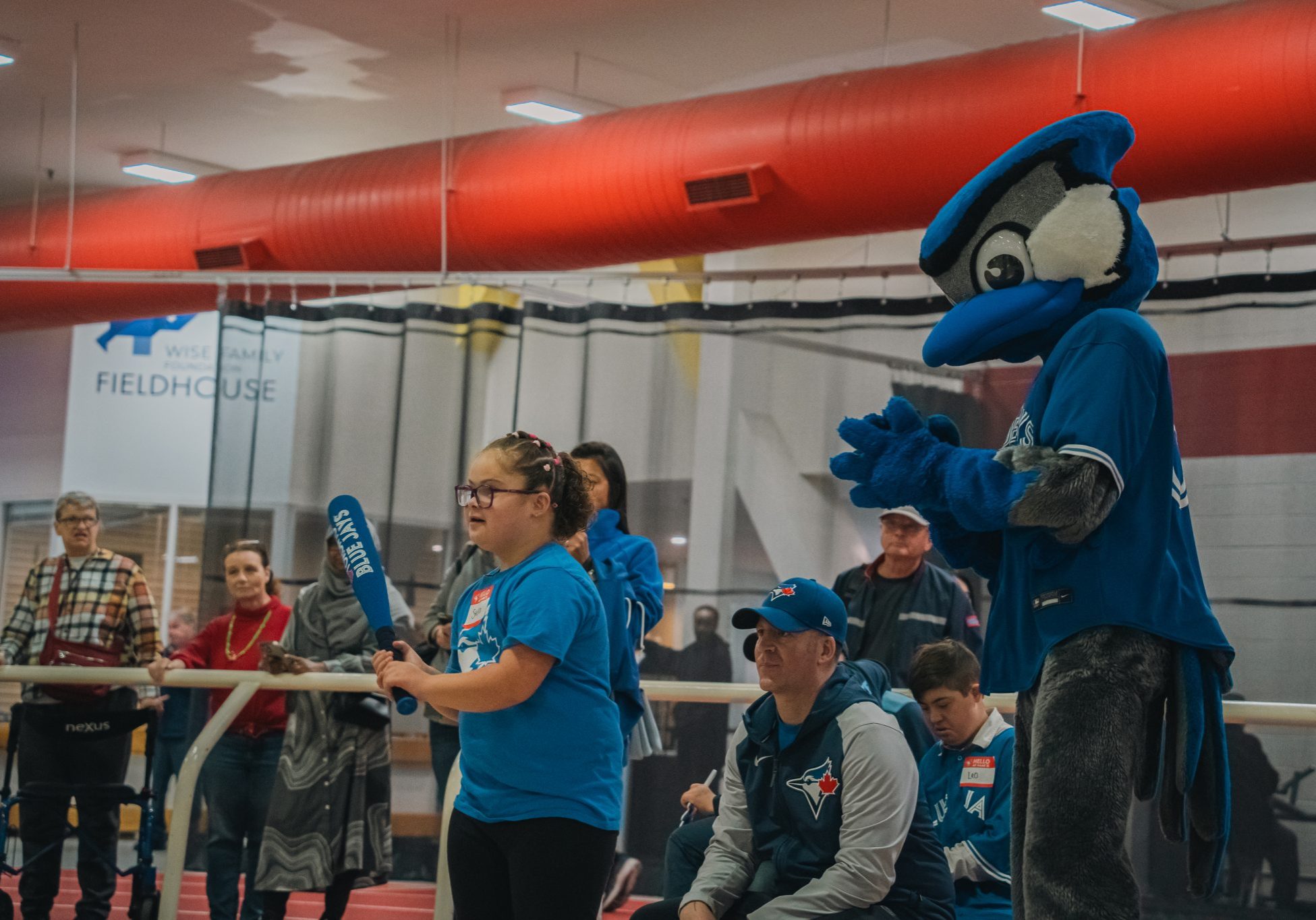 Child holds a foam baseball bat while the Toronto Blue Jays mascot, Ace, cheers from the sidelines during an indoor baseball activity.